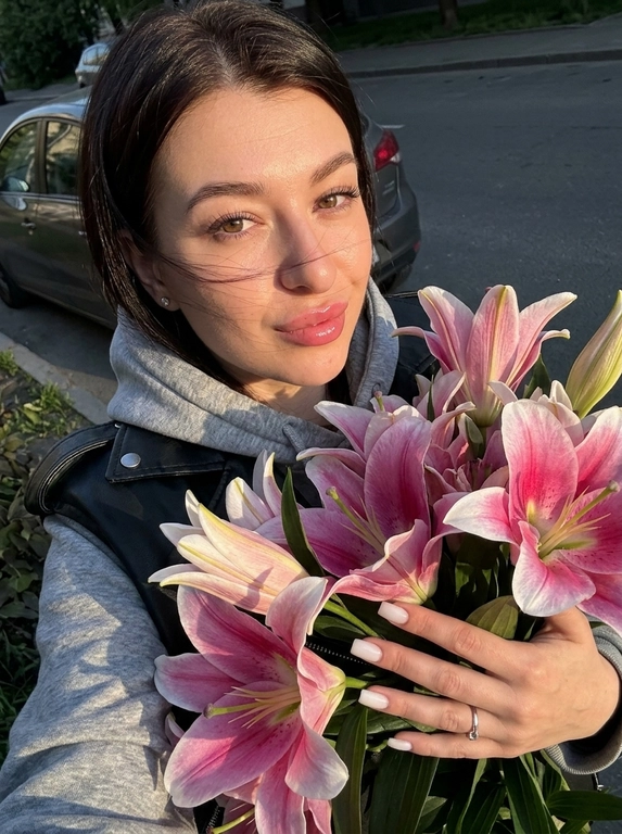 Используй лицо с моей референс-фотографии (face reference), сохрани сходство и узнаваемость на 100% — не менять овал лица, форму носа, бровей, губ, форму и цвет глаз, пропорции лица и тела. Создай реалистичное селфи на фронтальную камеру iPhone 17 Pro, телефон полностью находится за границей кадра. Девушка на улице возле припаркованной машины во время золотого часа. Кадр очень близкий: лицо занимает верхнюю половину фотографии, огромный букет поднимается снизу и частично закрывает свитер и куртку.
Свободной рукой девушка прижимает букет к груди и поддерживает его снизу.Камера расположена немного сверху и сбоку, девушка смотрит в камеру, подбородок слегка опущен. Выражение более игривое: глаза мягко смотрят в объектив, легкое кокетство.
Серое худи. Чёрная кожаная куртка свободно лежит на плечах. Огромные розово-белые лилии подходят практически к самому лицу; несколько лепестков находятся близко к объективу и слегка выпадают из глубины резкости.
Прическа —длинные прямые густые волосы с шелковистой поверхностью; ветер немного разбрасывает передние тонкие пряди по щеке и воротнику.
Макияж — особенно выразительные глаза: ухоженные  брови с хорошо различимыми волосками, чуть затемнённый внешний уголок, тонкая коричнево-чёрная межресничка. Ресницы длинные и воздушные, wispy, с чередованием тонких коротких и более длинных лучиков. Кожа ухоженная , на щеках свежий тёплый розово-персиковый румянец.
Губы  пухлые и чувственные естественные— juicy pink glass lips: мягкий натуральный розовый контур, середина чуть насыщеннее и холоднее, сверху плотный прозрачный блеск с влажными зеркальными бликами.
Маникюр — короткие молочный квалрат. На указательном пальце тонкое серебряное кольцо.
Свет: прямое заходящее солнце идёт сбоку и практически горизонтально. Оно интенсивно освещает лицо, кисть и ближайшие лепестки, формируя золотистые участки и довольно резкие тени. Тень от телефона/руки может слегка попасть на нижний край кадра — оставить её как реалистичную smartphone-деталь. В глазах маленькие яркие солнечные catchlights.
Фильтр: золотистые highlights, слегка бронзовая кожа, глубокие нейтральные тени, насыщенное синее небо. Pink/magenta цветов и губ немного усилены; зелень затемнена и десатурирована. Экспозиция слегка ниже автоматической, highlights сохранены, контраст высокий, но кожа не должна становиться чрезмерно резкой. Очень лёгкая смартфонная мягкость и естественная неоднородность экспозиции.  Букет огромный. Эффект живого кадра. Высокая детализация кожи, ткани,волос и ресниц . Легкий цифровой шум и зернисто