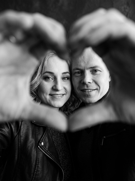 Ultra-realistic editorial photography of a smiling couple in a romantic and cinematic composition. The couple is centered in the background of the image, looking directly at the camera with a spontaneous, happy, and natural expression. In the foreground, extremely close to the lens and slightly out of focus, the couple’s hands form a heart shape perfectly framing their faces.

The composition creates strong visual depth, with sharp focus on the eyes and faces inside the heart formed by the hands. The hands occupy a large portion of the foreground, artistically blurred around the edges, creating a sophisticated cinematic effect.

Intimate, emotional, and elegant aesthetic inspired by modern perfume campaigns and premium lifestyle editorials. Soft studio lighting with delicate contrast and natural shadows. Minimalistic dark blurred background.

The couple wears minimalistic black leather jackets and sophisticated dark clothing. Genuine expressions, strong emotional connection, contemporary romantic atmosphere. Hyper-realistic skin texture, visible pores, detailed hair strands, authentic human appearance.

Black and white photography, rich tones, deep blacks, and soft highlights. 85mm lens, shallow depth of field, cinematic portrait photography, luxury editorial style, ultra detailed, hyper realistic, 8K.