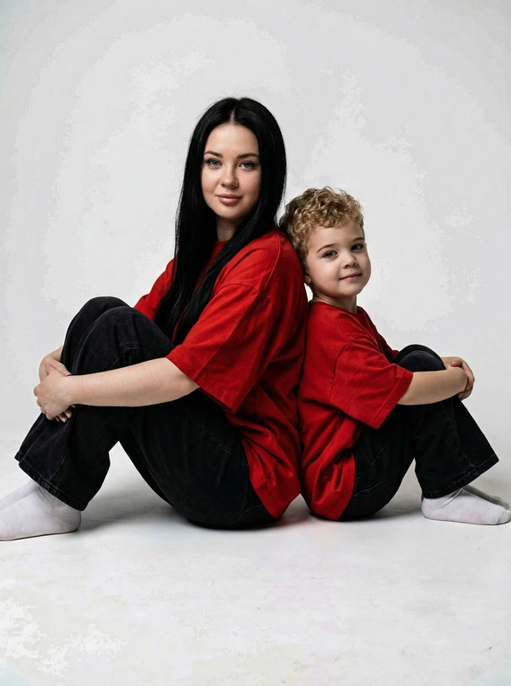 Гиперреалистичная фотография. Качество 8К. Используя портретное сходство с людьми с фотографий, создай фотографию: женщина в красной оверсайз свободной футболке и свободных черных джинсах, белых носках. Ребенок одет в красную оверсайз футболку, черные свободные джинсы и белые носочки. Женщина сидит на полу, согнув колени, смотрит прямо в кадр с еле заметной улыбкой. Сзади женщины в такой же позе сидит ребенок облокотившись спиной к спине женщины, с легкой улыбкой. Темный стиль модного редакционного журнала, глубокие тени и драматическое студийное освещение, белый фон, элегантная и мощная композиция, ультрареалистичный, высокая детализация, художественный портрет.