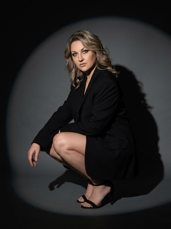 Модная студийная съёмка в стиле fashion editorial. Не менять внешность женщины. Женщина одета в только в чёрный оверсайз пиджак. Профессиональный макияж с акцентом на кожу и глаза. Один направленный прожектор формирует чёткое круглое световое пятно на темно-сером бесшовном гладком фоне и выразительную тень за спиной. Контрастный кинематографичный свет, глубокие тени, драматичный минимализм. Атмосфера — уверенность, сила, эстетика high fashion. высокая детализация кожи, реалистичная текстура, съёмка уровня модного журнала. Средний план. Женщина сидит на корточках, одна рука лежит на колене, корпус слегка наклонён вперёд. Уверенный прямой взгляд в камеру. На ногах чёрные босоножки на тонком каблуке. Чёткая тень позади усиливает драматизм кадра.

Какая будет прическа?: Волосы распущены, уложены в легкие волны.