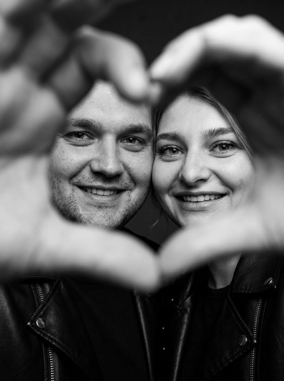 Ultra-realistic editorial photography of a smiling couple in a romantic and cinematic composition. The couple is centered in the background of the image, looking directly at the camera with a spontaneous, happy, and natural expression. In the foreground, extremely close to the lens and slightly out of focus, the couple’s hands form a heart shape perfectly framing their faces.

The composition creates strong visual depth, with sharp focus on the eyes and faces inside the heart formed by the hands. The hands occupy a large portion of the foreground, artistically blurred around the edges, creating a sophisticated cinematic effect.

Intimate, emotional, and elegant aesthetic inspired by modern perfume campaigns and premium lifestyle editorials. Soft studio lighting with delicate contrast and natural shadows. Minimalistic dark blurred background.

The couple wears minimalistic black leather jackets and sophisticated dark clothing. Genuine expressions, strong emotional connection, contemporary romantic atmosphere. Hyper-realistic skin texture, visible pores, detailed hair strands, authentic human appearance.

Black and white photography, rich tones, deep blacks, and soft highlights. 85mm lens, shallow depth of field, cinematic portrait photography, luxury editorial style, ultra detailed, hyper realistic, 8K.