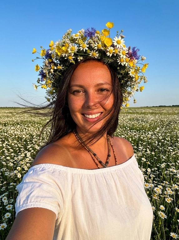 Женщина делает селфи. На ней белое легкое платье с открытыми плечами, лёгкое хлопковое  платье. Кожа загорелая. У нее на голове объёмные цветочные венки из мелких полевых ромашек . Венки должны быть пышные, натуральные, свежие, как летний flower crown. Цветы расположены вокруг головы, без искусственного вида.
Её волосы развиваются от ветра слегка падая на лицо.
Фон: летнее поле с ромашками. Открытое небо без облаков слегка голубое.
Не размывай фон. Голова слегка наклонена к плечу.
Рук в кадре нет.
Горизонт прямой , волосы сильно развиваются от ветра. Солнечная погода в золотой час.