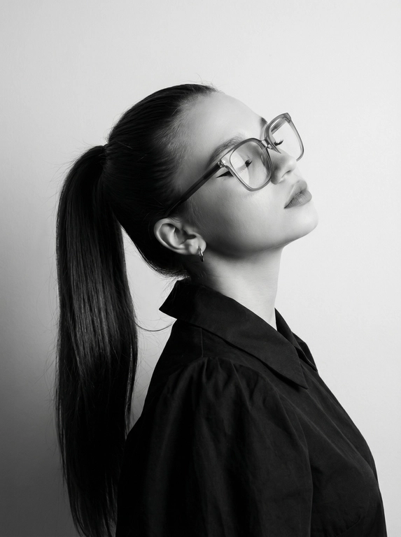 Чёрно-белый минималистичный фотореалистичный портрет, студийный мягкий свет спереди‑сверху, нейтральная светлая однотонная стена как фон. Девушка в профиль: голова запрокинута назад, подбородок поднят, глаза закрыты — расслабленная fashion-поза с акцентом на линии шеи и контуры лица. Обязательно точно сохранить черты лица с референс-фото (без искажений); крупные квадратные очки в массивной полу-прозрачной оправе с естественными бликами. Детали лица: длинные ресницы и чёткие стрелки, выразительные губы, естественная текстура кожи, аккуратный ретуш. Одежда: тёмный объёмный верх с выразительным воротником, маленькая серьга-кольцо. Стиль: высокая графичность и контраст, чистые линии, лёгкая драматичность, максимальная детализация и фотореализм, аккуратные светотеневые переходы, подчёркнутые контуры.

Какая будет прическа?: Гладкие волосы, высокий длинный густой хвост.