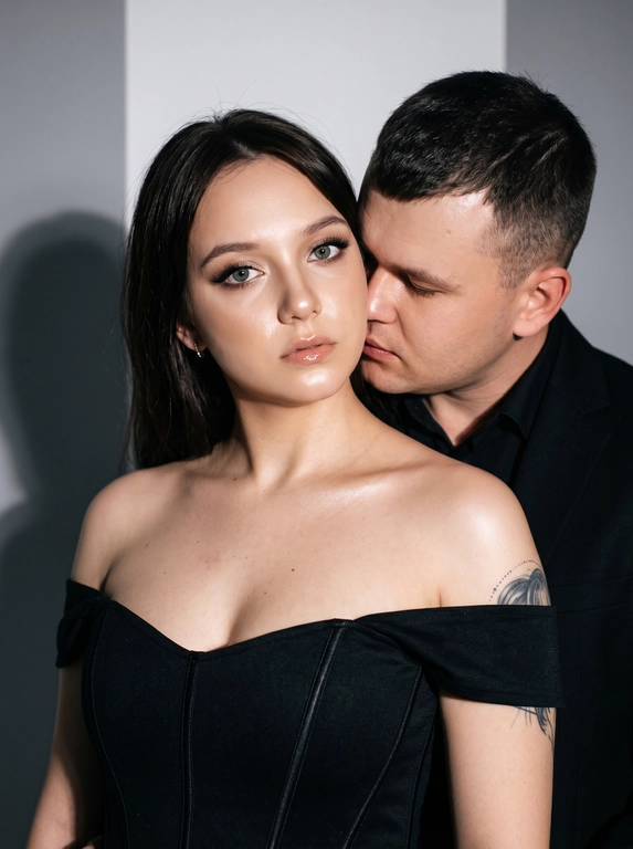 Не меняя черты лица, студийная fashion-съемка пары в минималистичном интерьере с серо-белым фоном, атмосфера гламура и интимной близости, ракурс — крупный портрет по грудь с легким поворотом корпуса, женщина на переднем плане, мужчина позади склоняется к её плечу, поза — она слегка отвернута от камеры с прямым взглядом, он касается лицом её шеи/плеча, одежда — на женщине черный корсет/бандо с открытыми плечами, на мужчине классический черный пиджак и рубашка, макияж — glam (ровный сияющий тон, сильный хайлайтер, четкий контуринг, стрелки, густые ресницы, глянцевые губы), свет — жесткая фронтальная вспышка + мягкий заполняющий свет, выраженные тени позади, эффект flash photography / editorial, холодно-нейтральная цветокоррекция с контрастом и глянцем кожи, высокая детализация, slight film grain, shallow depth of field, съемка на Canon EOS R / 50mm, f/2.0, direct flash, studio look.