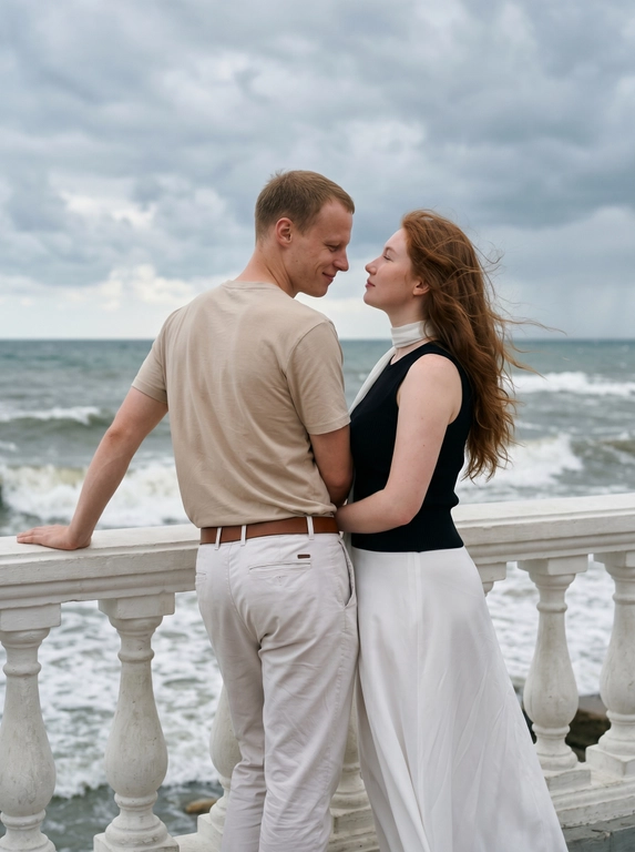 Пара стоит у белого каменного балюстрадного ограждения с видом на море. Мужчина слева, спиной к камере, корпус в 3/4 вправо, левая рука опирается на перила, голова повернута вправо к женщине, смотрит на неё с нежностью. Женщина справа, корпус в 3/4 влево, стоит очень близко к мужчине, смотрит ему в лицо снизу вверх. Одежда: мужчина — светлая бежевая футболка с коротким рукавом, светлые брюки, коричневый ремень; женщина — длинное платье с чёрным облегающим верхом без рукавов и светлой струящейся юбкой до пола, белый тонкий шарф на шее. Волосы слегка развеваются. Фон: море, пасмурное небо. Мягкий естественный свет, 50mm, лёгкое зерно.