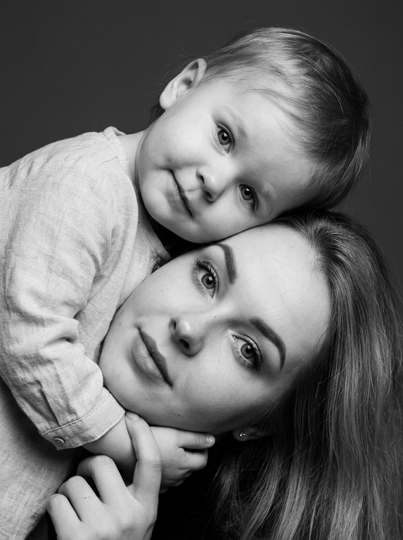 Родитель и ребёнок с внешностью как на приложенных фото, художественная семейная студийная фотосессия. Композиция — вертикальный портрет, крупный план лиц. Родитель внизу смотрит прямо в камеру, лицо фронтально. Ребёнок сверху, голова повернута набок, щека лежит на голове родителя, смотрит в камеру с нежной улыбкой. Рука ребёнка обнимает родителя за шею, видна в кадре, родитель держит руку ребёнка своей рукой.
Одежда: ребёнок в белой льняной рубашке, родитель в чёрной водолазке. 
Фон — тёмно-серый почти чёрный однотонный студийный, создаёт контраст. Освещение — мягкое боковое студийное, создаёт объём и глубину.
Чёрно-белая художественная фотография с выразительными контрастами. Атмосфера нежная, интимная, спокойная.