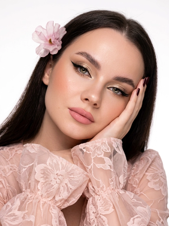На фотографии запечатлена женщина крупным планом, позирующая для портрета. Снимок подчеркивает её ухоженную внешность и стильный образ.
Лицо и выражение:
•  Глаза:  ярко выделены макияжем. На верхних веках нанесены мерцающие золотисто-бежевые тени, дополненные четкими, длинными черными стрелками («кошачий глаз»). Ресницы густые и длинные, подчеркивающие глубину взгляда.
•  Брови: Густые, уложенные вверх (эффект ламинирования), что придает лицу современный и открытый вид.
•  Кожа: Тон кожи ровный, с нежным персиковым румянцем на скулах. Кончик носа и скулы слегка подсвечены хайлайтером для придания лицу сияния.
•  Губы: Выразительные, накрашены матовой помадой нежно-розового оттенка. Верхняя губа имеет четкий контур.
•  Поза: Женщина слегка наклонила голову вправо и подперла щеку правой рукой. Этот жест придает снимку нотку нежности и кокетства.Маникюр темно-бордовый,квадратный.
Одежда:
•  Ткань украшена принтом в крупный кружевной цветок .
•  Рукава имеют пышные воланы или оборки у запястий, что добавляет образу романтичности и женственности.
Фон и освещение:
•  Фон белый студийный
•  Освещение мягкое, направленное на лицо, что позволяет рассмотреть все детали макияжа и создает приятную, эстетичную картинку.
Общее впечатление:
Фотография представляет собой классический бьюти-портрет. Образ женщины сочетает в себе элегантность, современность и романтичность. Тщательно проработанный макияж и стильная одежда создают гармоничный и привлекательный облик.

Какого цвета будет платье?: На женщине надета блузка или платье из легкой, полупрозрачной ткани нежно розового цвета.

Какого цвета будет цветок?: В волосах один нежный ,воздушный розовый цветок.
