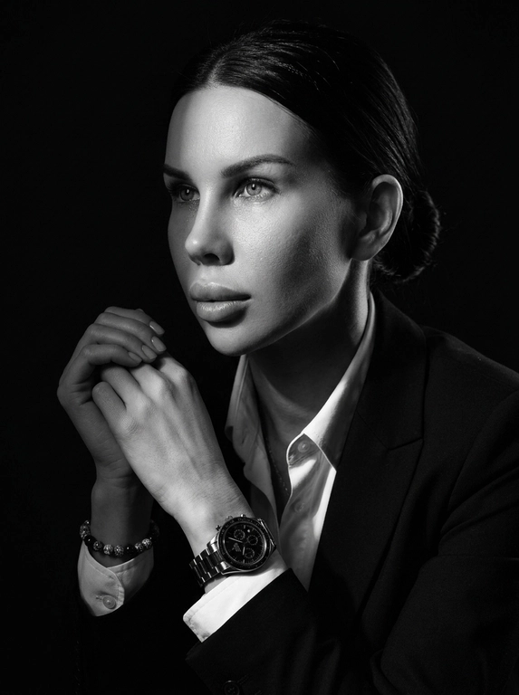 A hyper-realistic 8K cinematic portrait of a uploaded person,
captured in a dramatic Chiaroscuro style. The subject is seated at a
three-quarter angle, leaning slightly forward with a relaxed yet
commanding posture. His face is turned away from the lens, with
one side of his face clearly illuminated while the other is
completely lost in deep, velvety black shadows. His expression is
contemplative, not looking at the camera. The pose is precise: his
hands are clasped together near his chest; one hand is held
straight with fingers slightly tilted, while the other hand is
positioned so only the fingers are visible, resting over the first. He
wears a luxury black chronograph watch with a detailed metal link
bracelet, and a beaded bracelet. His attire is a sharp
black suit jacket over a white dress shirt with the top buttons
open, showing refined fabric textures. The background is a solid,
seamless black. The lighting is sharp and directional, highlighting
the intricate details of his hair,
the moisture in his eyes, and the glint off the watch face.
Cinematic black-and-white color grade with rich contrast and
smooth, natural skin transitions. Shot on 85mm lens, f/1.8, high-
resolution commercial photography

Доп. характеристики: Черно белая фотография , максимально реалистичное лицо, легкий макияж,  волосы в низком пучке