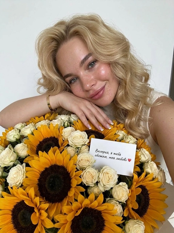 На фотографии запечатлена женщина, которая держит в руках очень большой и пышный букет цветов.
Её кожа сияет, загорелого оттенка, на губах глянцевый блеск, улыбка лёгкая.

она положила руку и голову на букет, взгляд в камеру и находится позади букета.
 Букет: Это объемная композиция, состоящая из контрастного сочетания двух видов цветов:
    – Подсолнухи: Крупные, яркие желтые подсолнухи с темной сердцевиной, которые создают акцент в букете.
    – Розы: Множество нежных кремово-белых роз (похожих на кустовые), которые заполняют пространство между подсолнухами, придавая композиции мягкость и элегантность.Среди цветов видна небольшая подарочная карточка или визитка.

Она одета в вязаный топ с высоким горлом, без рукавов, свет молочный. Маникюр аккуратный, Френч, квадратная форма, на запястье браслет Cartier. Композиция выглядит очень ярко, позитивно и празднично благодаря сочетанию солнечных оттенков подсолнухов и нежных тонов роз.

Фон - белая стена. Высокая детализация кожи. Живое фото, без ретуши на лице. Мобильная съемка. Лёгкая пленка, зерно. Raw. ультрареалистичный дорогой лайфстайл снимок.

Доп. характеристики: Не меняй черты лица. Строго сохрани внешность! Визитке надпись: Валерия, я тебя обожаю, моя любовь ❤️