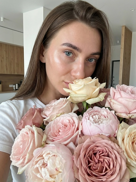     
An ultra-realistic close-up portrait of a young woman, partially obscured by a huge bouquet of roses, against the backdrop of a modernly renovated apartment. The frame is cropped from the forehead to the middle of the chest, so that her face and flowers take up almost the entire image. 
 
She has smooth skin with a natural glow; highlighter accentuates the upper cheeks and nose, giving them a dewy sheen. Her eyes are clear and very expressive, framed by thick, dark lashes and softly shaped, naturally arched brows. She looks directly at the camera over the flowers, creating intimate eye contact. 
 
Her hair is thick and straight, parted slightly off-center. It falls freely back from her face, with a touch of volume at the crown. She wears a simple white T-shirt, revealing only the neckline and a small portion of her shoulder, adding a minimalist touch and focusing attention on her face and the flowers. 
 
In front of her, she holds a large, lush bouquet of garden roses and peonies, so that the flowers cover almost everything from her nose down. Roses in varying shades of pale pink, soft pink, and cream, with some petals slightly tinted to a darker dusty pink at the edges. The flowers are fully open, with abundant petals and a wavy, romantic texture; some outer petals display delicate wrinkles and slight imperfections, enhancing the natural realism. Small patches of green stems and leaves peek through, but the composition is dominated by the soft cloud of petals. 
 
A single cream rose is positioned directly beneath her nose, as if she were gently inhaling its fragrance. The bouquet is exquisitely detailed: subtle color transitions, veining on the petals, and the way the petals catch the light, creating tiny highlights and shadows within each bloom are visible. 
 
The lighting is soft and even, with minimal shadows, as if the photo was taken with diffused natural light or a softbox. This highlights the model's skin and makes the pastel colors soft and gentle. The background gives the image a fresh, clean look—romantic, minimalist, and high-resolution.
