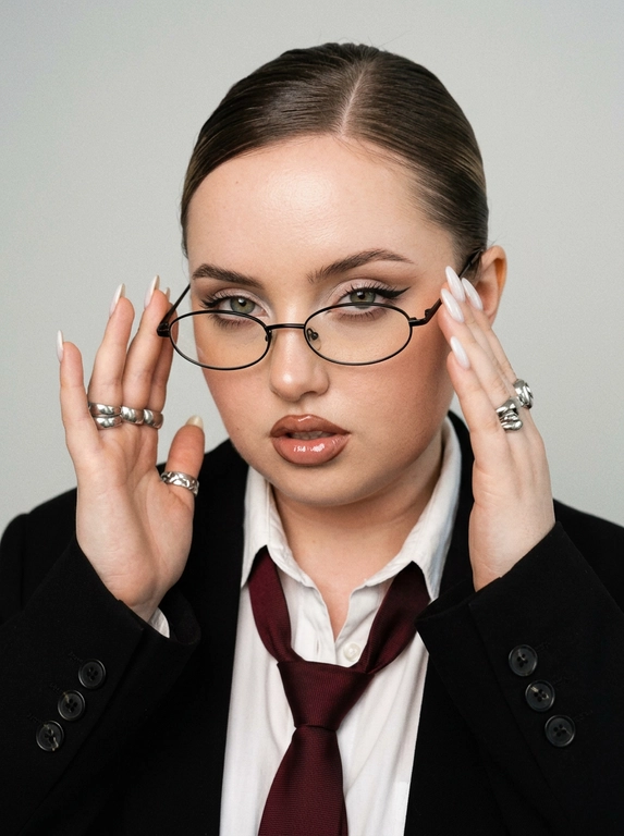 Используй приложенное фото и создай снимок, где крупным планом изображена эта женщина, одетая в белую рубашку с расстегнутыми верхними пуговицами, черный оверсайз пиджак и расслабленно завазанный бордовый мужской галстук, что создает деловой, но неформальный образ. Черты лица, телосложение, цвет волос остаются неизменными с референсом. Волосы убраны назад в гладкий пучок, боковой пробор. На лице женщины узкие овальные очки в тонкой черной оправе. Выражение лица уверенное, надменное, смотрит в камеру из под очков и поправляет очки двумя руками. Губы слегка приоткрыты. На пальцах видны массивные серебрянные кольца.  Маникюр. длинные ногти миндальной формы, покрытые молочным лаком. Макияж в стиле офисной сирены: тонкие брови, выразительные стрелки на глазах, растушеванные светлые тени, губы подведены коричневым карандашом, глянцевые. Освещение мягкое и равномерное, создающее студийную атмосферу. Фон чистый, однотонный, светло-серый, который контрастируют с темным пиджаком и галстуком. Общая атмосфера снимка - сексуальная, стильная и современная. Y2K стиль, зернистость, легкий винтажный фильтр.