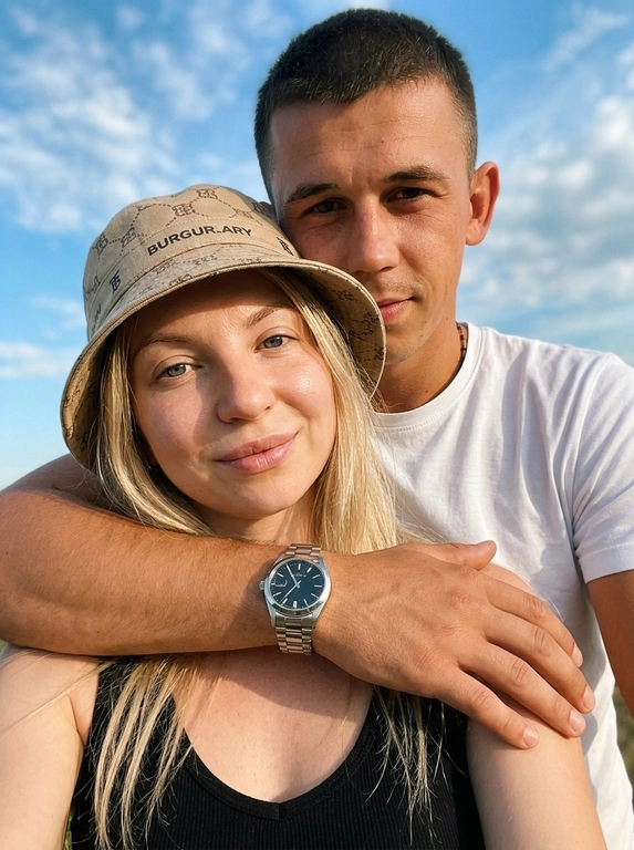 Кинематографичный кадр крупным планом (selfie style), пара в объятиях (классическое "обнимашки сзади") на фоне ярко-голубого неба с облаками.  Женщина на ней бежевая панама с брендовой надписью. Её кожа выглядит сияющей, подсвеченной мягким естественным солнечным светом. Выражение лица спокойное, глубокий взгляд, направленный прямо в камеру. Мужчина стоит строго позади девушки и обнимает её сзади, одна его рука обнимает её за плечи/шею спереди — предплечье лежит поперёк её ключиц, кисть свободно свисает с её плеча, на запястье классические серебряные часы с черным циферблатом. Мужчина в белой футболке, выражение лица доброжелательное, его взгляд прямо в объектив. Ракурс сверху, камера сфокусирована на лице девушки. Высокий контраст, насыщенные цвета, мягкая прорисовка деталей кожи. Эффект пленочной фотографии, легкое зерно, атмосфера летнего дня, эстетика социальных сетей. Телефона не видно в кадре.