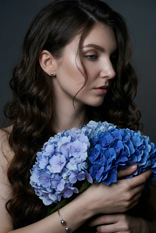 Создайте ультрареалистичный портрет, не меняя черты лица, крупным планом 3/4, с мягкой, освещённой кожей, атласной отделкой и естественным блеском, где женщина изображена в профиле. Спокойное и чувственное выражение лица, губы слегка полуоткрыты. Она носит серебряные серьги-шары и простой серебряный браслет на запястье.
Фон нейтральный, в светло-сером тоне, с тёмным, целенаправленным низким угловым освещением, которое подчёркивает контур лица, текстуру кожи и объём волос. Стиль редактирования — высокая мода, чёткий фокус 8K, реалистичная текстура, изысканная и элегантная атмосфера.


Длинные волнистые волосы покрывают часть лица объёмными локонами, выпадающими на лицо и плечо. 

Цветы: Держит в руках огромный букет голубых гортензий
