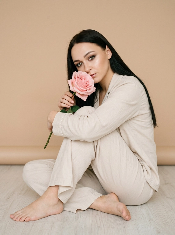Сохрани внешность человека с исходного фото. Создай студийное фото на котором человек сидит на полу обнимая колени одной рукой. Второй рукой человек держит красивую нежную розу с большим бутоном, бутон цветка закрывает часть её лица. Макияж красивый профессионалный, ровная кожа с естественным сиянием, немного хайлайтера, дымчатые тени, красивые тонкие графичные стрелки, губы в прозрачном блеске. Одежда: светлый хлопковый костюм по типу пижамы, рубашка с длинным рукавом и свободные брюки. Но ноги босые. Фон бежевый, бесшовный и однотонный, фокус на человеке. Естественный свет допустимо лёгкое зерно. Высокое качество фото.

Прическа: Волосы ухоженные уложены прямо