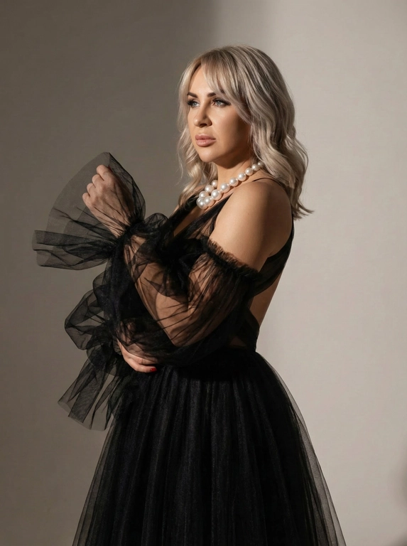   Не меняя внешности женщины с фото, перед нами предстает изящная фигура, подчеркнутая изысканным вечерним образом, где главная роль отведена сложному платью с пышными рукавами и объемным подолом из черного тюля. Женщина стоит в полуобороте, ее тело слегка изгибается, создавая динамичную линию, которая придает образу живость и легкую драму. Верхняя часть платья практически открыта, подчеркивая плечи и тонкую талию, при этом прозрачные, многослойные воздушные воланы вокруг рук словно парят в пространстве, создавая атмосферу таинственности и высокой моды. Волосы светлве до плеч уложенные волнами, чёлка шторка дополняя элегантность образа и открывая лицо, на котором сосредоточено мягкое световое пятно, подчеркивающее выразительный макияж с акцентом на глаза и губы. Взгляд женщины направлен в сторону, и кажется, что она задумчиво смотрит в даль, о чем-то мечтая или размышляя, что добавляет кадру интригующий эмоциональный подтекст. На шее заметно массивное украшение из белых жемчужин, контрастирующее с темным нарядом и подчеркивающее изысканный вкус. Освещение построено таким образом, что один бок модели окутан мягким светом, создающим объем и глубину, а другой частично скрыт в тенях, что подчеркивает текстуру и прозрачность многочисленных слоев ткани. Задний фон минималистичен и нейтрален, позволяя сосредоточиться на фигуре и одежде модели, при этом игра света и тени оживляет пространство вокруг, добавляя эффектную кинематографичность всему образу. Вся композиция напоминает кадр из стильного модного фильма, где каждая деталь тщательно продумана и несет смысл, создавая ощущение утонченности и драматизма, фотореалистичное изображение, высокая текстурная детализация, высокое качество, разрешение 8K.