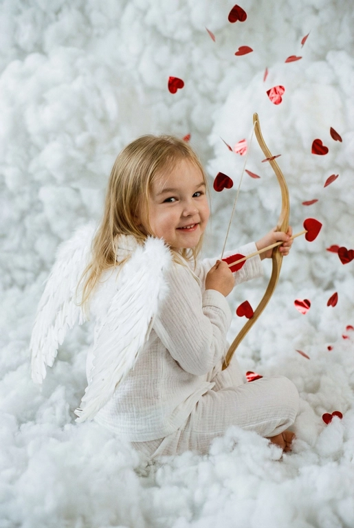 Использовать реальные черты лица человека с референса, естественную текстуру кожи и настоящий цвет и длину волос. Личность должна оставаться неизменной.

Y2K. Сказочная сцена с купидоном, сфотографированная как реальный момент.
Человек сидит среди плотных, пушистых белых облаков, которые полностью заполняют передний, средний и задний план, образуя бесшовную белую среду с многослойной глубиной.

Поза вполоборота: корпус слегка повернут вбок, одно плечо выдвинуто вперед, как будто человек натягивает и отпускает небольшой лук с красной стрелой в форме сердца; руки подняты с легким напряжением, указывающим на движение при выстреле.

Несмотря на поворот тела, голова повернута к объективу, взгляд направлен прямо в камеру. Выражение лица радостное и игривое, с мягкой улыбкой.

Наряд — простая белая пижама: свободная рубашка и брюки из муслина.
На спине — крылья из белых перьев, настоящие и фактурные, с четкими бликами по краям каждого пера.

Вокруг объекта в воздухе хаотично летают только конфетти в форме сердечек — маленькие красные сердечки из глянцевой фольги, расположенные на разном расстоянии: часть в переднем плане, часть растворяется среди облаков.

Без рассеянного света, без мягкого свечения, без студийного глоу.
Текстура кожи реалистичная и детализированная, без сглаживания и без фильтров.
Тонкая зернистость, волшебное, но искреннее ощущение Y2K-редакции — выглядит как фотография, а не визуализация.