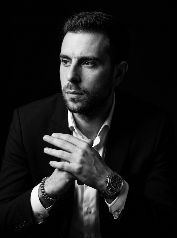 A hyper-realistic 8K cinematic portrait of a uploaded person,
captured in a dramatic Chiaroscuro style. The subject is seated at a
three-quarter angle, leaning slightly forward with a relaxed yet
commanding posture. His face is turned away from the lens, with
one side of his face clearly illuminated while the other is
completely lost in deep, velvety black shadows. His expression is
contemplative, not looking at the camera. The pose is precise: his
hands are clasped together near his chest; one hand is held
straight with fingers slightly tilted, while the other hand is
positioned so only the fingers are visible, resting over the first. He
wears a luxury black chronograph watch with a detailed metal link
bracelet, and a beaded bracelet. His attire is a sharp
black suit jacket over a white dress shirt with the top buttons
open, showing refined fabric textures. The background is a solid,
seamless black. The lighting is sharp and directional, highlighting
the intricate details of his hair,
the moisture in his eyes, and the glint off the watch face.
Cinematic black-and-white color grade with rich contrast and
smooth, natural skin transitions. Shot on 85mm lens, f/1.8, high-
resolution commercial photography

Доп. характеристики: Не меняя внешность , сделай красиво