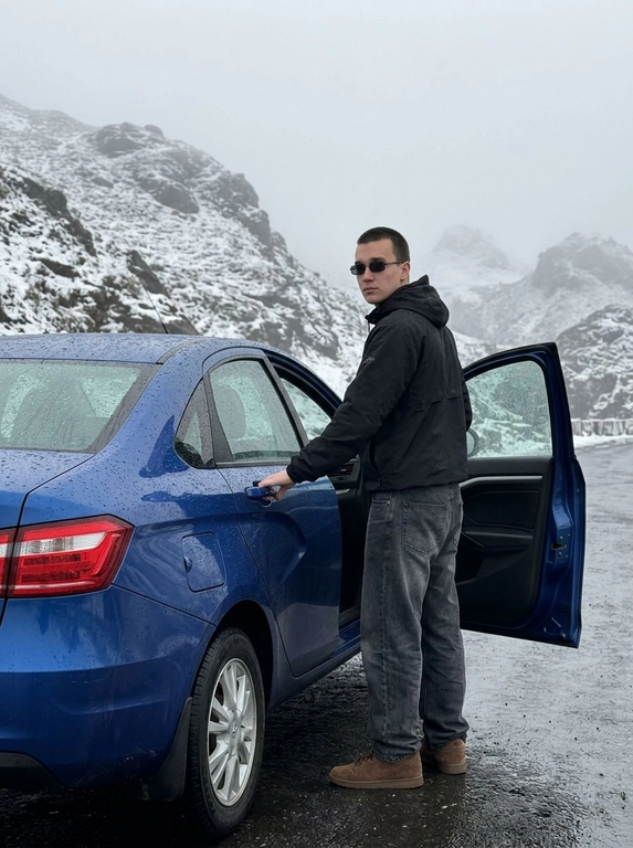 Не менять черты лица человека с исходного фото. Человек рядом с стоит рядом с машиной, машина на горной дороге, дорога мокрая от тумана, машина в каплях от тумана. Чёрный анорак Planta Rosa, маленькие чёрные солнцезащитные очки без оправы, мешковатые серо-чёрные джинсы с низкой посадкой, длинные, коричневые замшевые кроссовки Nike Dunk, руки в карманах. Машина без номеров. Фон — гиперреалистичные заснеженные холмы Мегхалайи, без зелени, туманные горы, пасмурный мягкий дневной свет, без размытия, без изменения лица или позы, без дополнительных объектов. Фото снято на iPhone 17 Pro, без фильтров на лице, без ретуши, перспектива снизу, формат 9:16, машина как на исходном фото, фон не размывать, притемнённая экспозиция, открывает дверь машины, стоит спиной, смотрит в камеру, будто сейчас сядет за руль.


Какая будет машина?: Синяя Lada Vesta